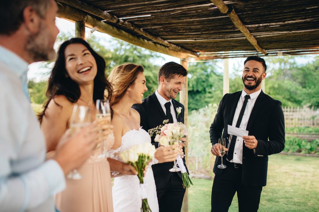 Best man smiling while giving toast to bride, groom, and friends