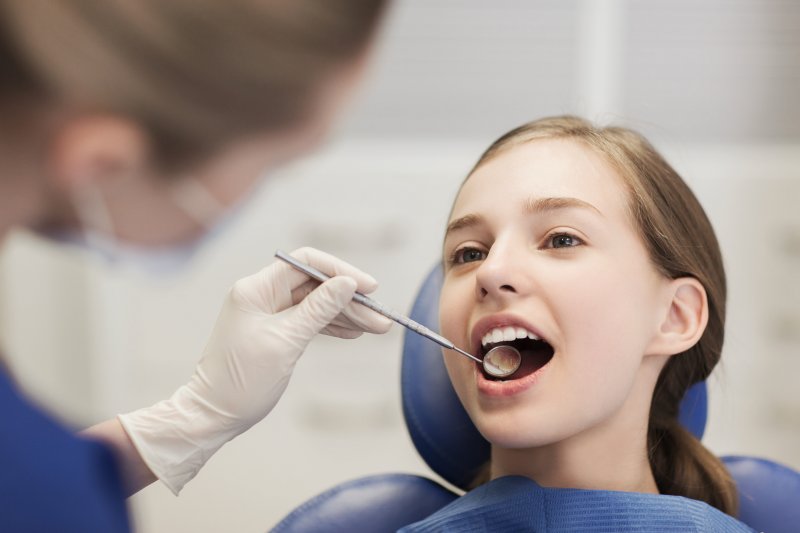 Girl in dental chair opening her mouth for dentist holding dental mirror in gloved hand