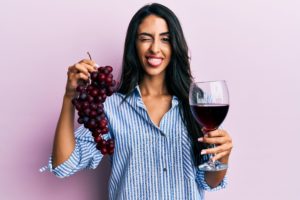 Woman in striped shirt holding a glass of wine in one hand and red grapes in the other while smiling and winking at the camera